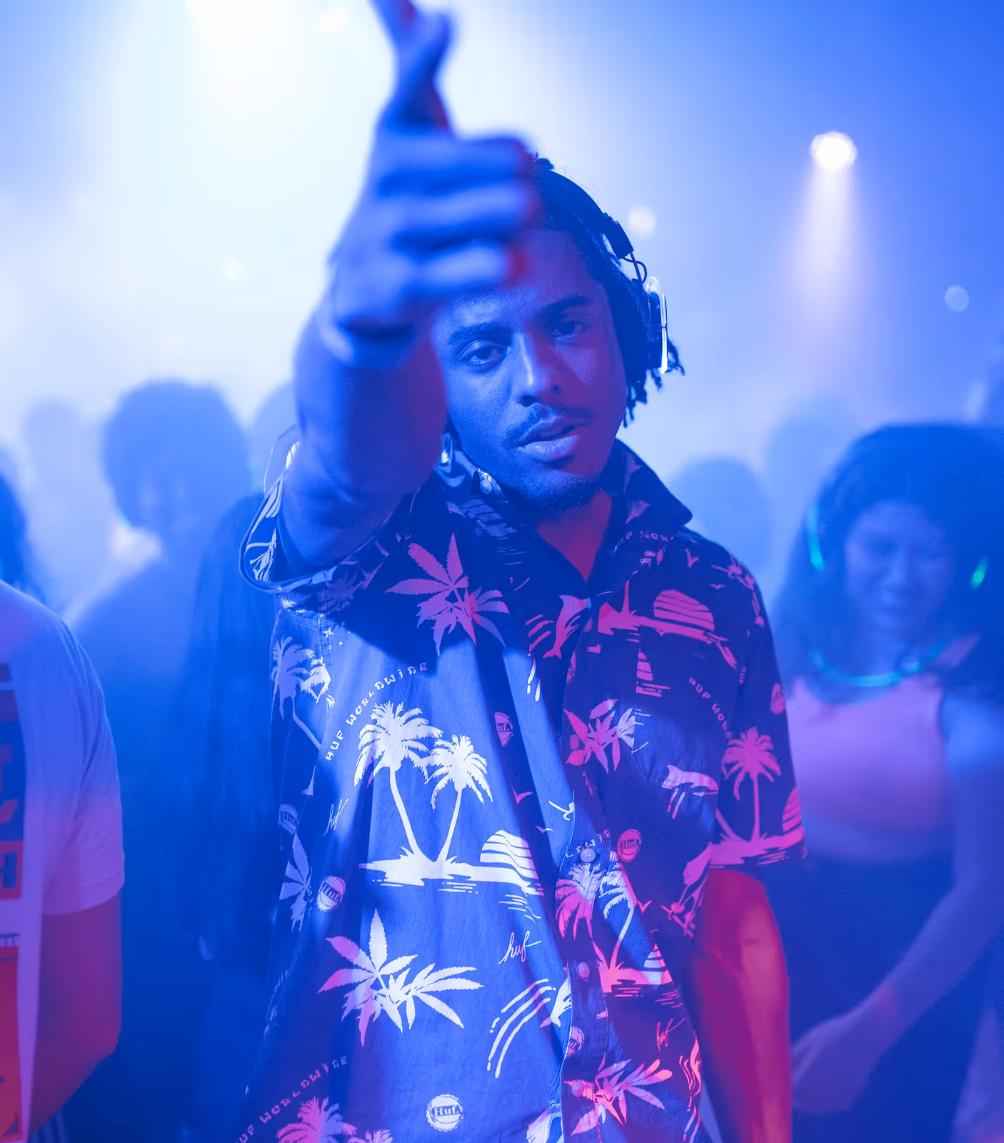 Man met koptelefoon en Hawaiiaans overhemd geeft een duim omhoog op een feest met blauwe en paarse verlichting.