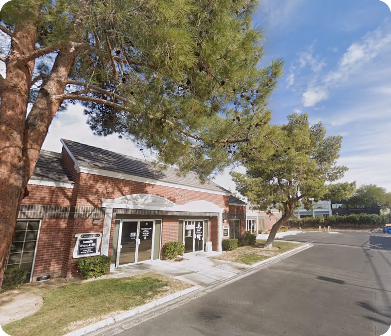 Brick office building with glass doors, surrounded by trees and a paved parking area under a partly cloudy sky.