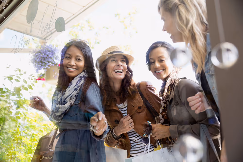Four women smiling and looking through a shop window while holding shopping bags.