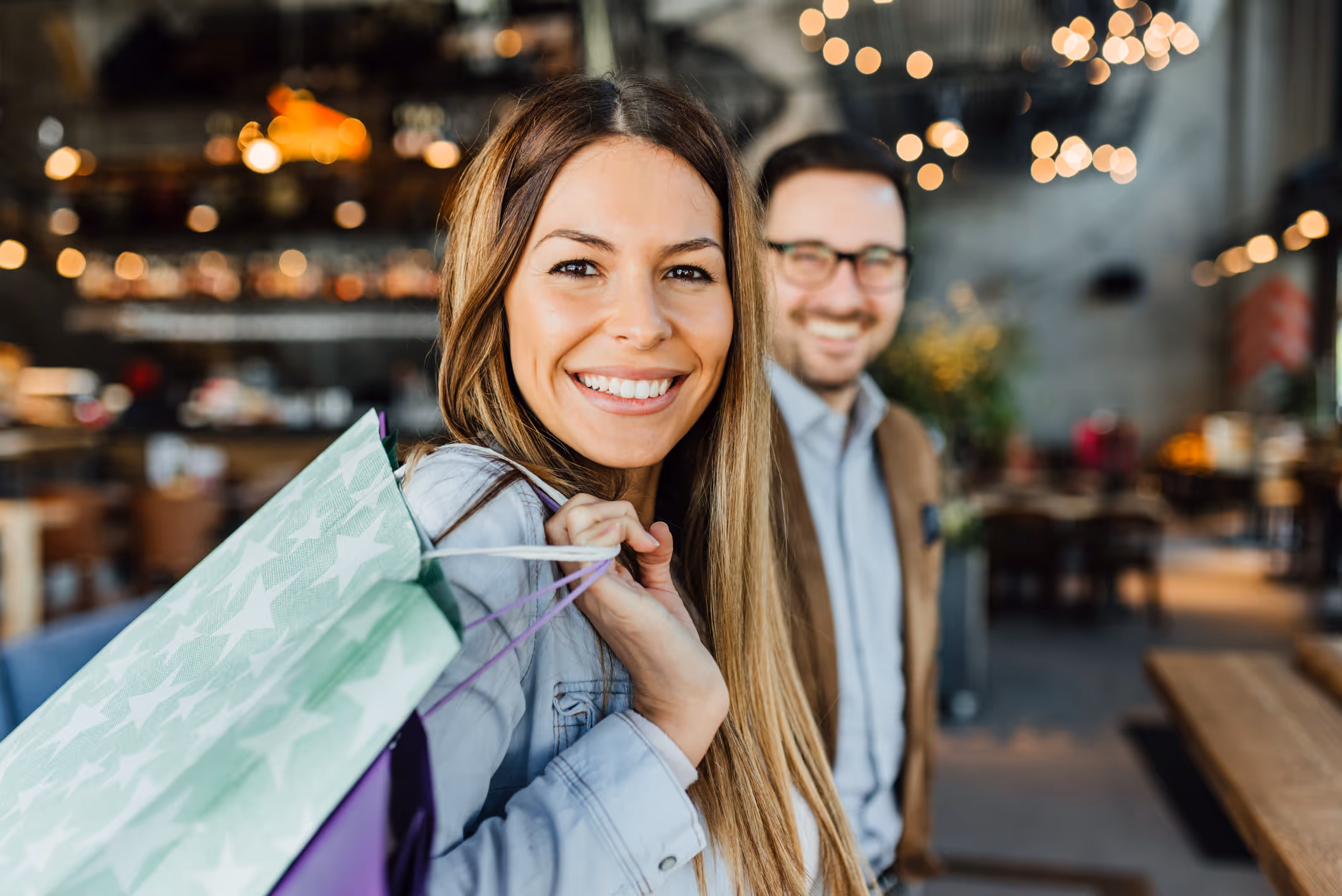 Smiling woman holding shopping bags over her shoulder with a blurred man in the background inside a warmly lit indoor space.
