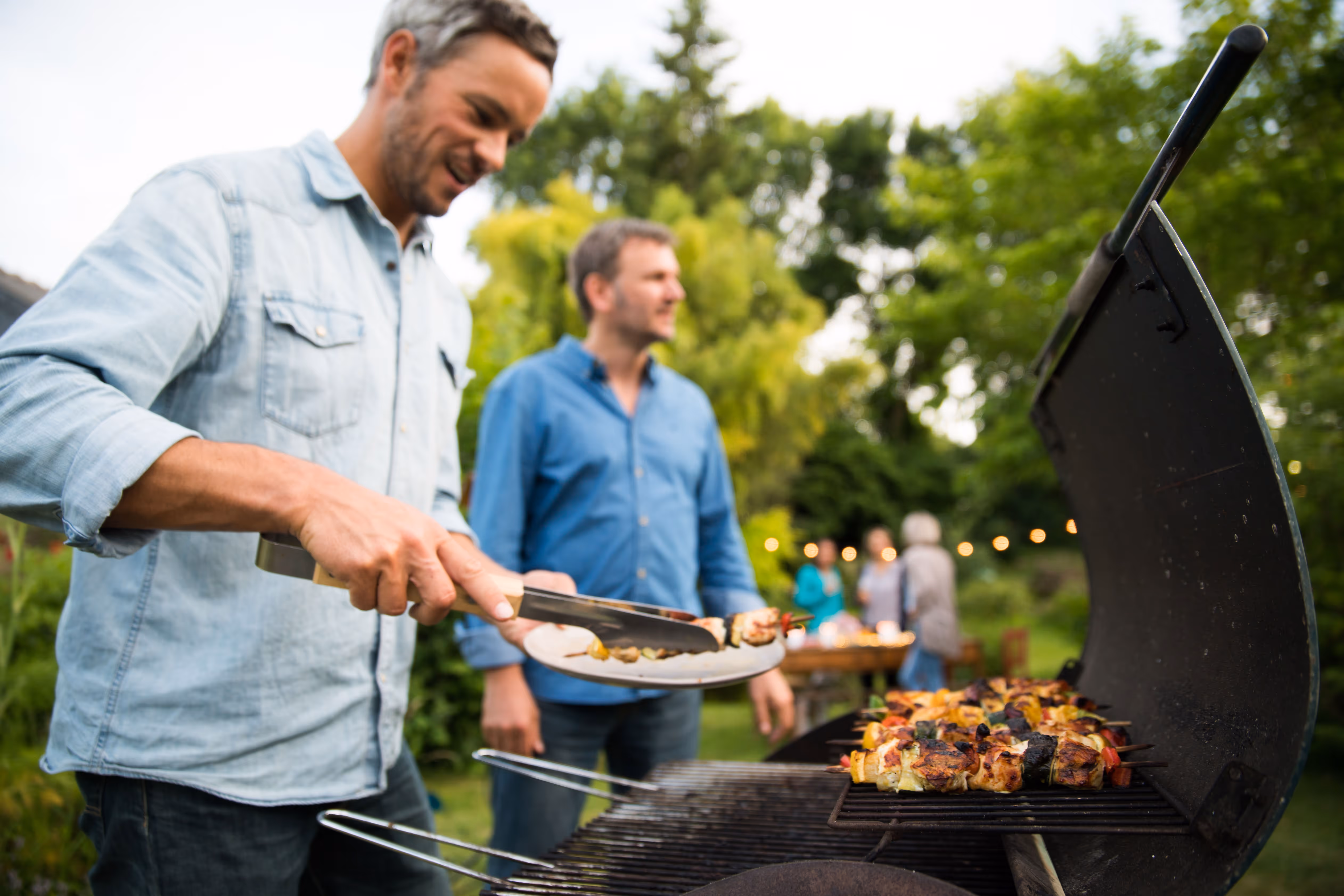 Man grilling skewers with vegetables and meat outdoors during a backyard gathering with people in the background.