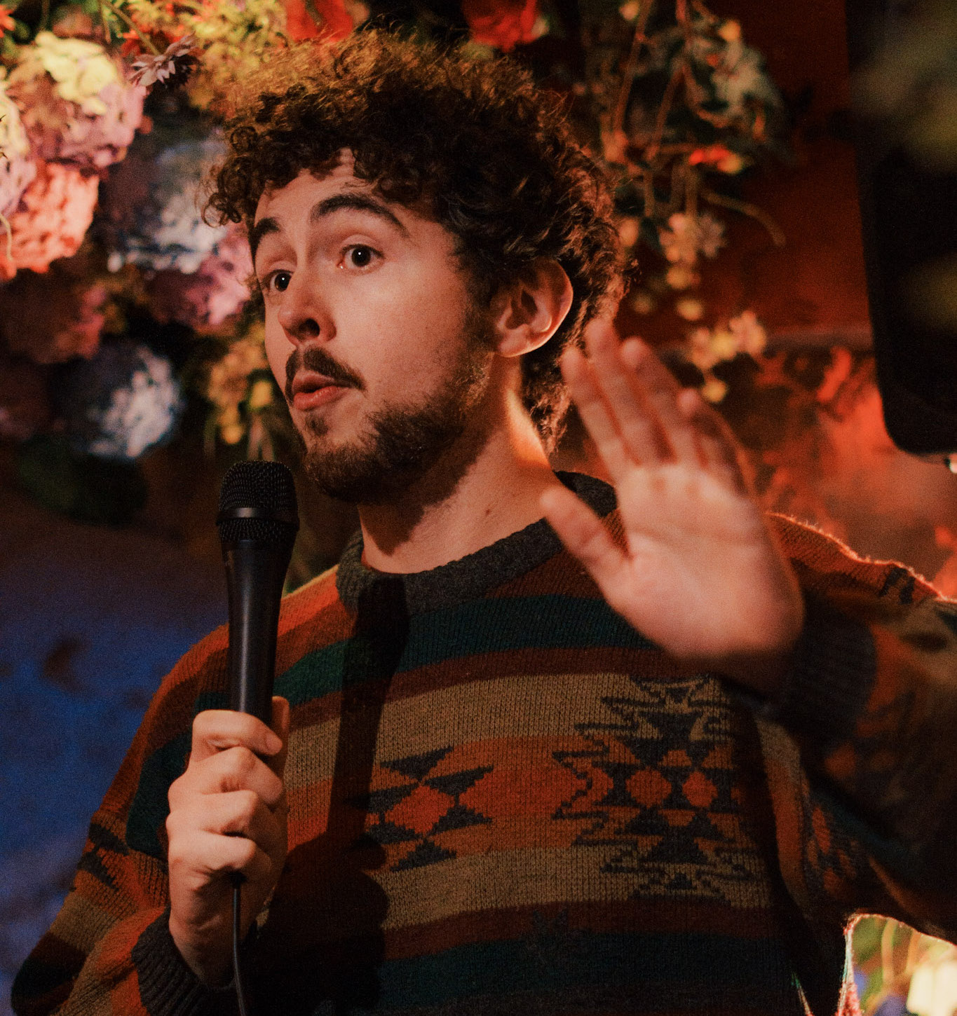 Man in a patterned sweater speaking into a microphone with his hand raised, against a floral and warm-lit background.