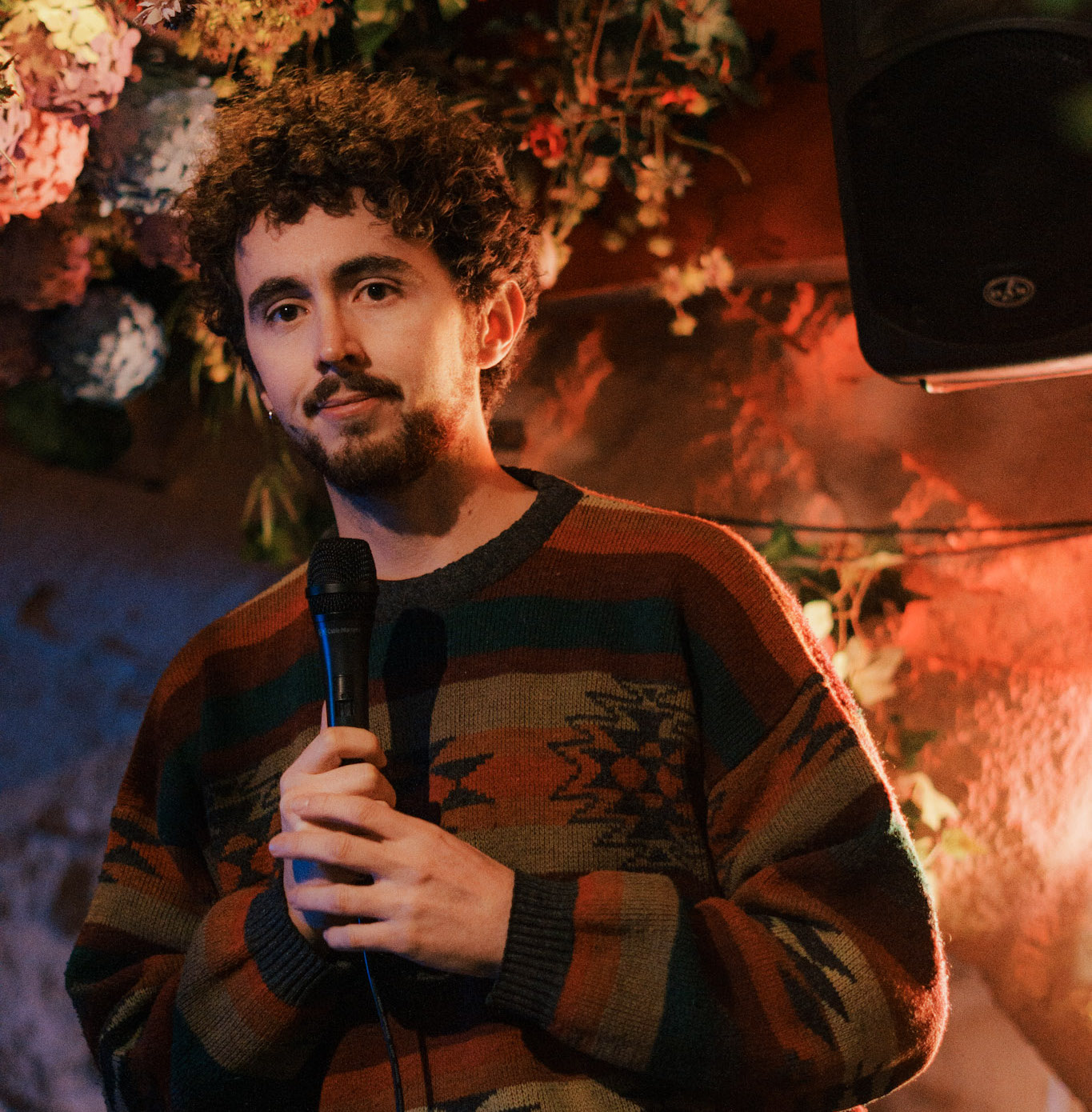 Man with curly hair and beard holding a microphone, standing in a warmly lit room with floral decorations in the background.