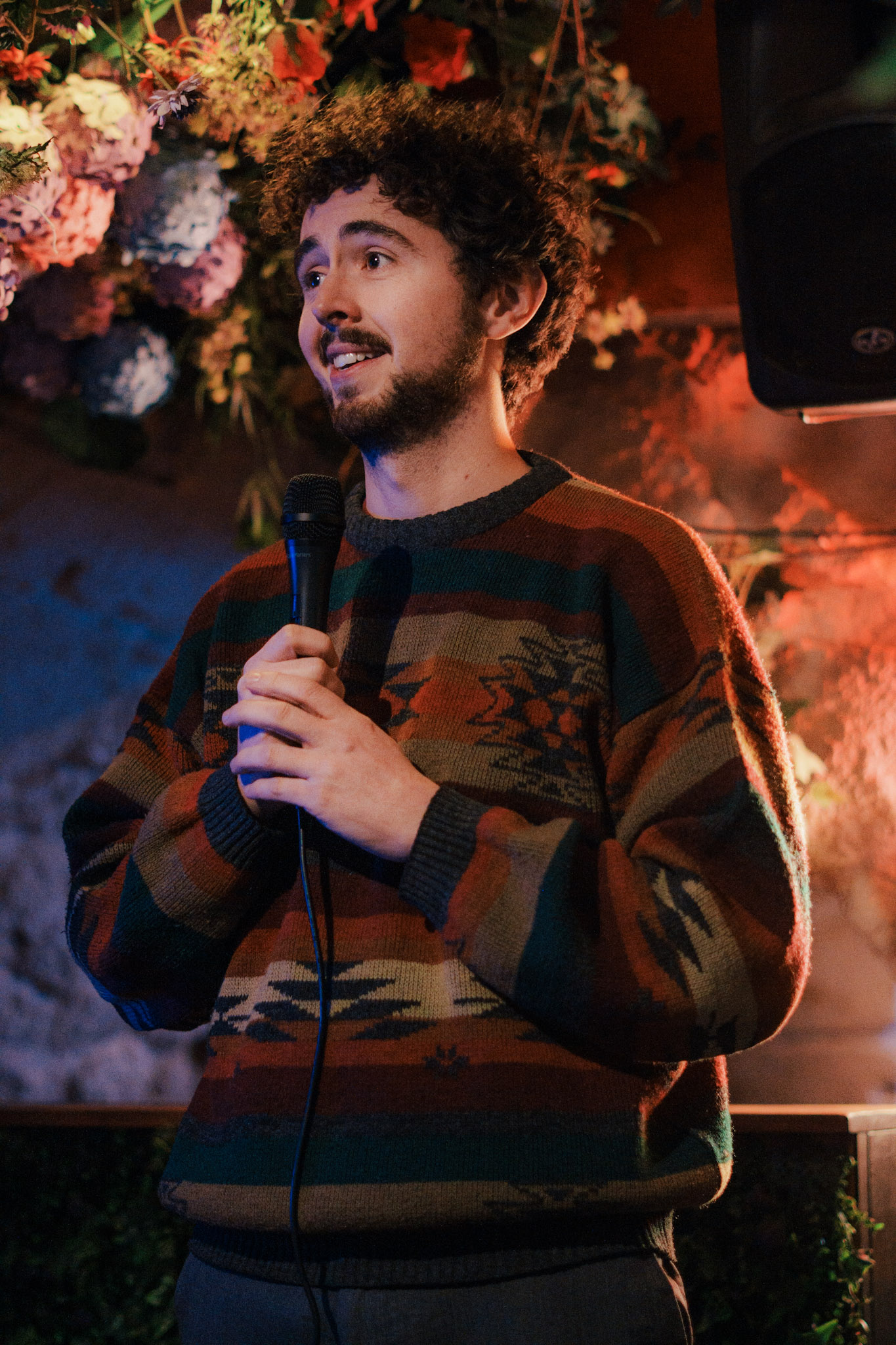 Young man with curly hair and beard wearing a patterned sweater, holding a microphone and speaking on stage with colorful floral decorations in the background.