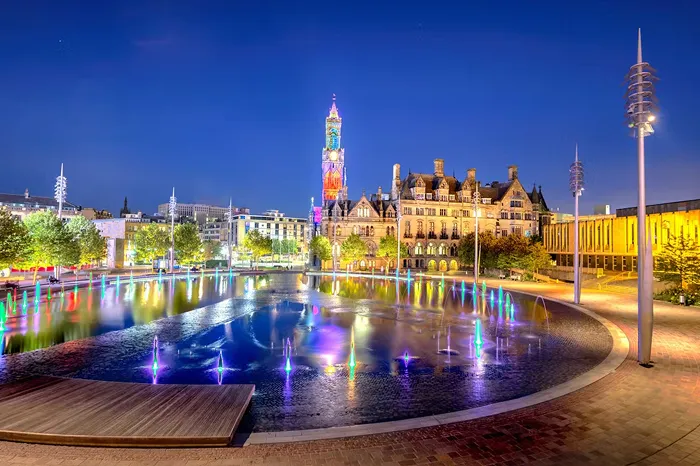 Night view of a colorful illuminated fountain in front of a historic building with a tall lit clock tower.