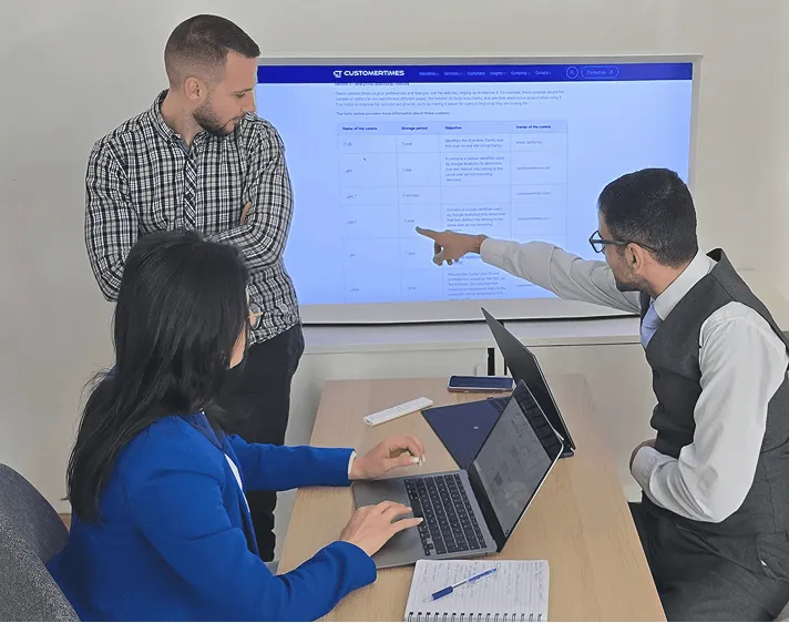 Three colleagues in a meeting room discussing data displayed on a large screen, one man is pointing at the screen while a woman works on a laptop.