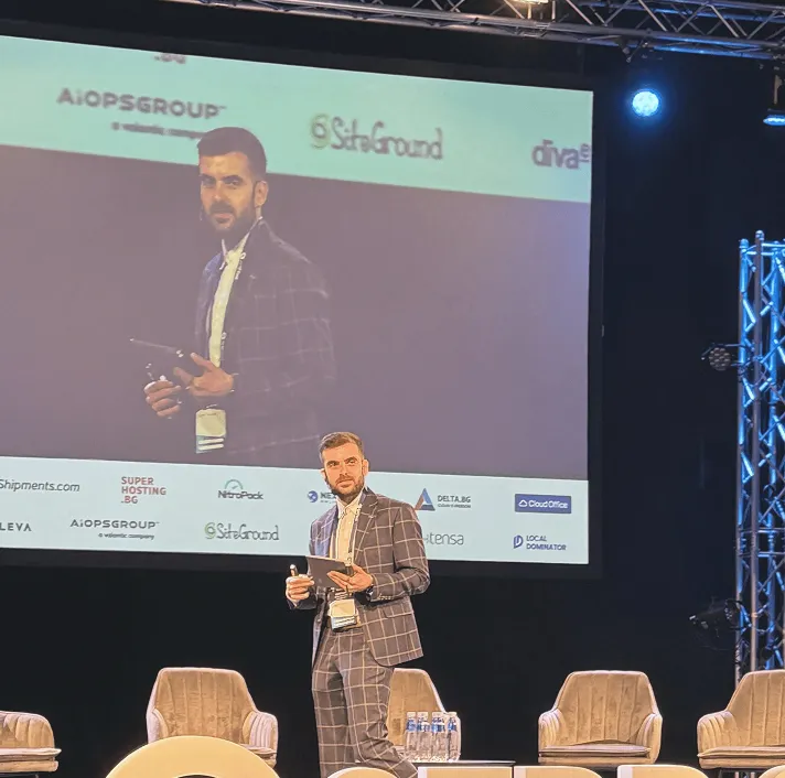 Man in a checkered suit holding a tablet and microphone, standing on stage with chairs and water bottles, with his image projected on a large screen behind him.