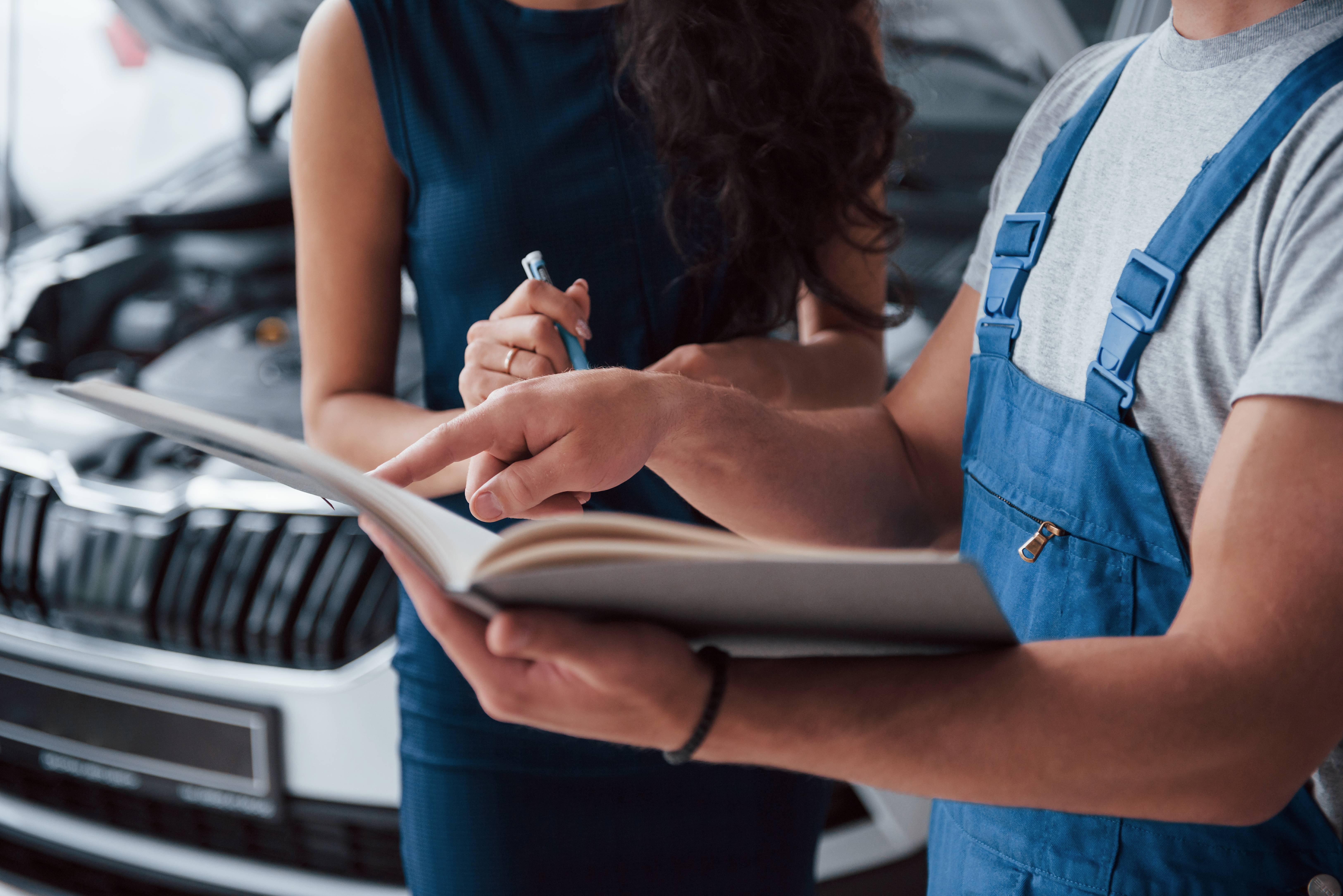 Mechanic in blue overalls showing a book to a woman holding a pen near a car with an open hood.