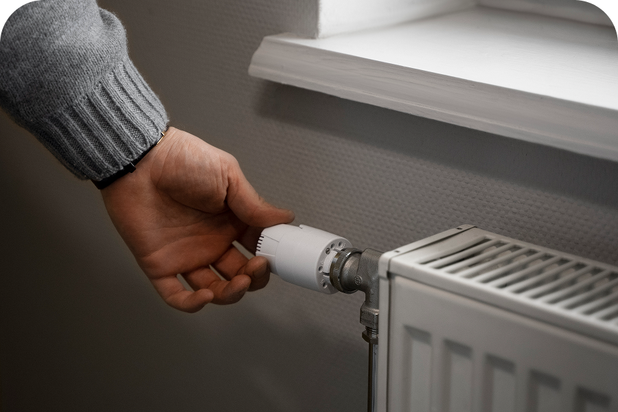 Hand adjusting the thermostat valve on a white radiator mounted below a window.