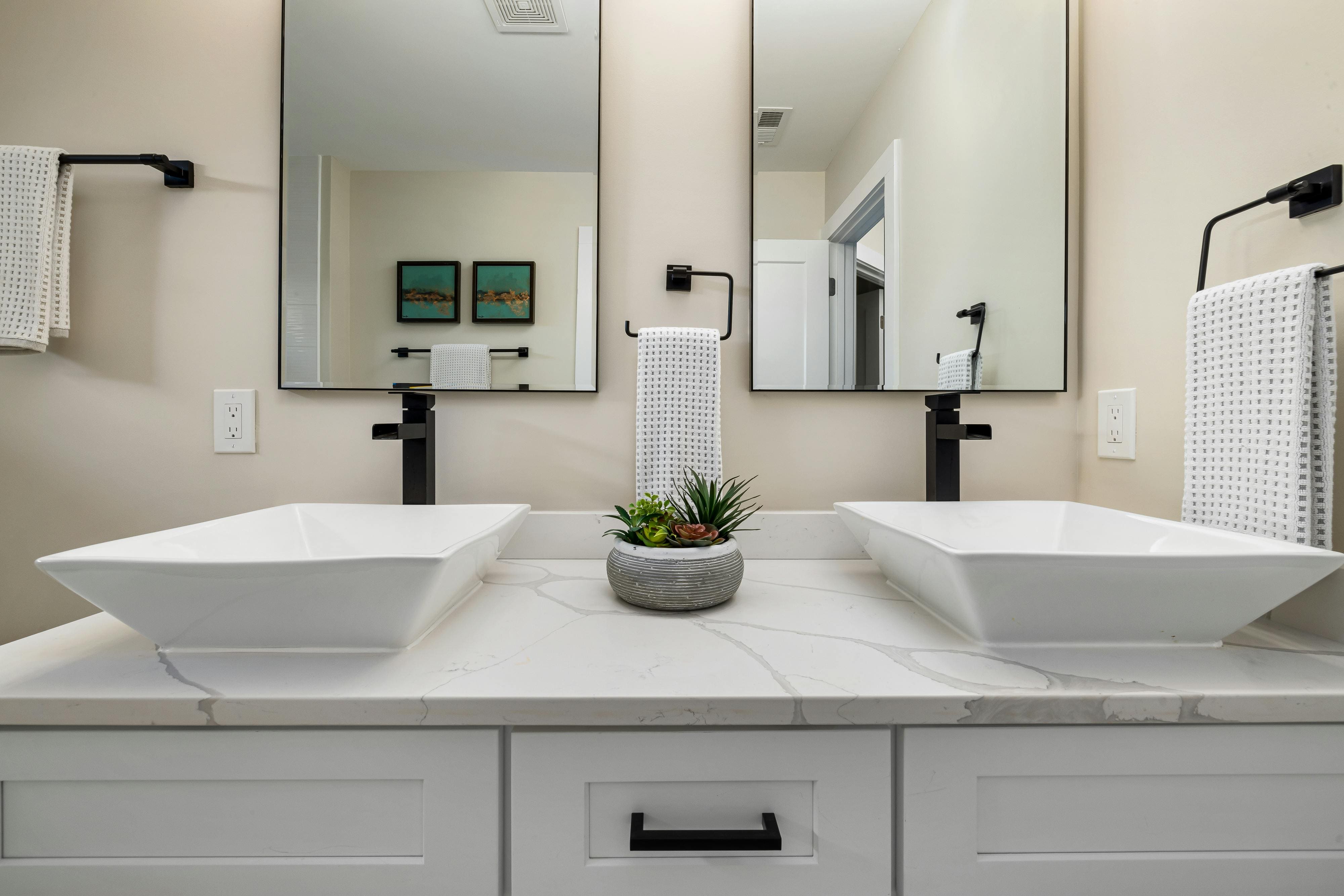 Modern bathroom vanity with dual white rectangular vessel sinks, black faucets, two mirrors, and a small succulent plant on the marble countertop.