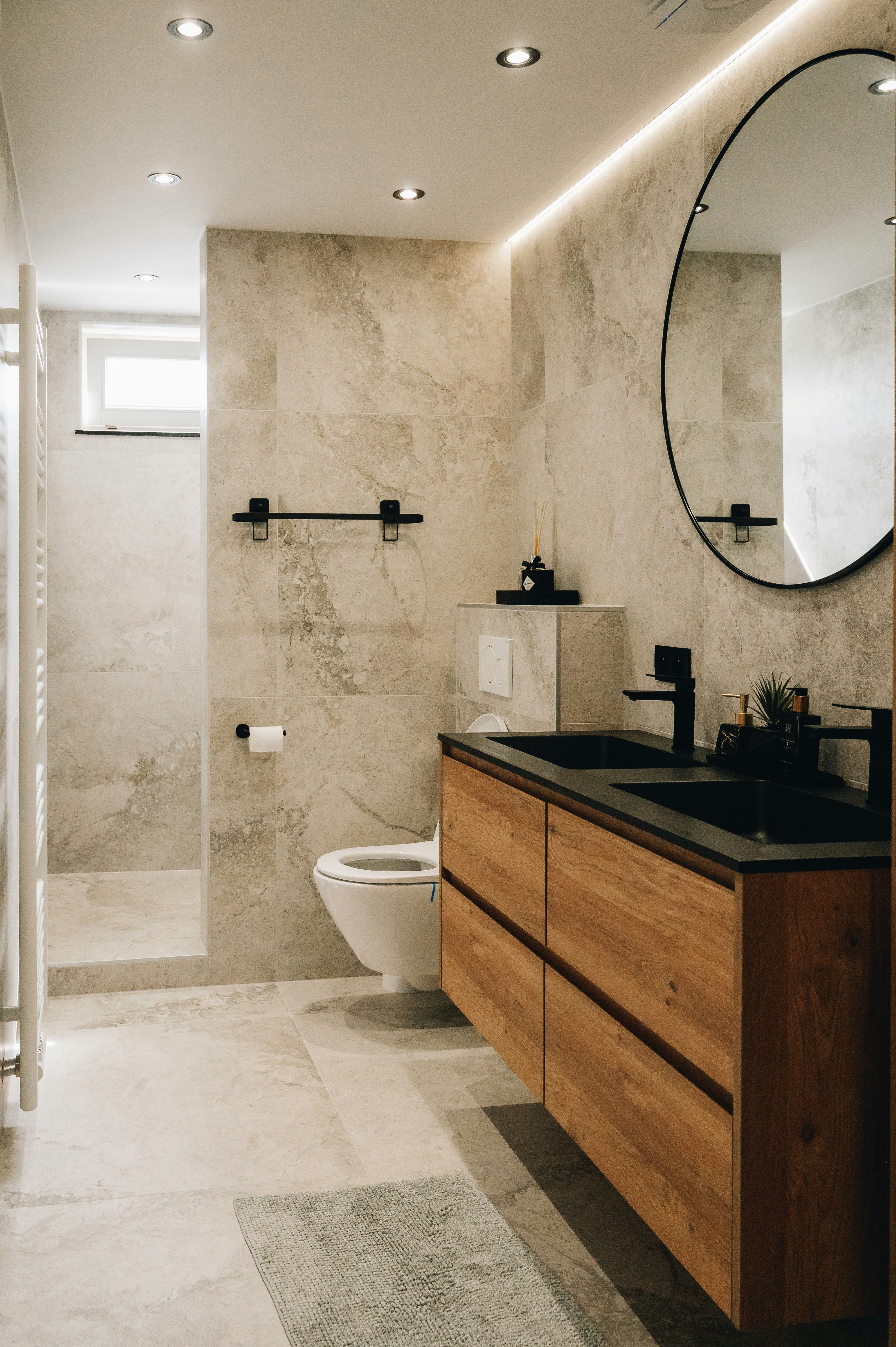 Modern bathroom with beige marble walls and floors, a wall-mounted toilet, walk-in shower, wooden vanity with black countertop and double sinks, and a large round mirror.
