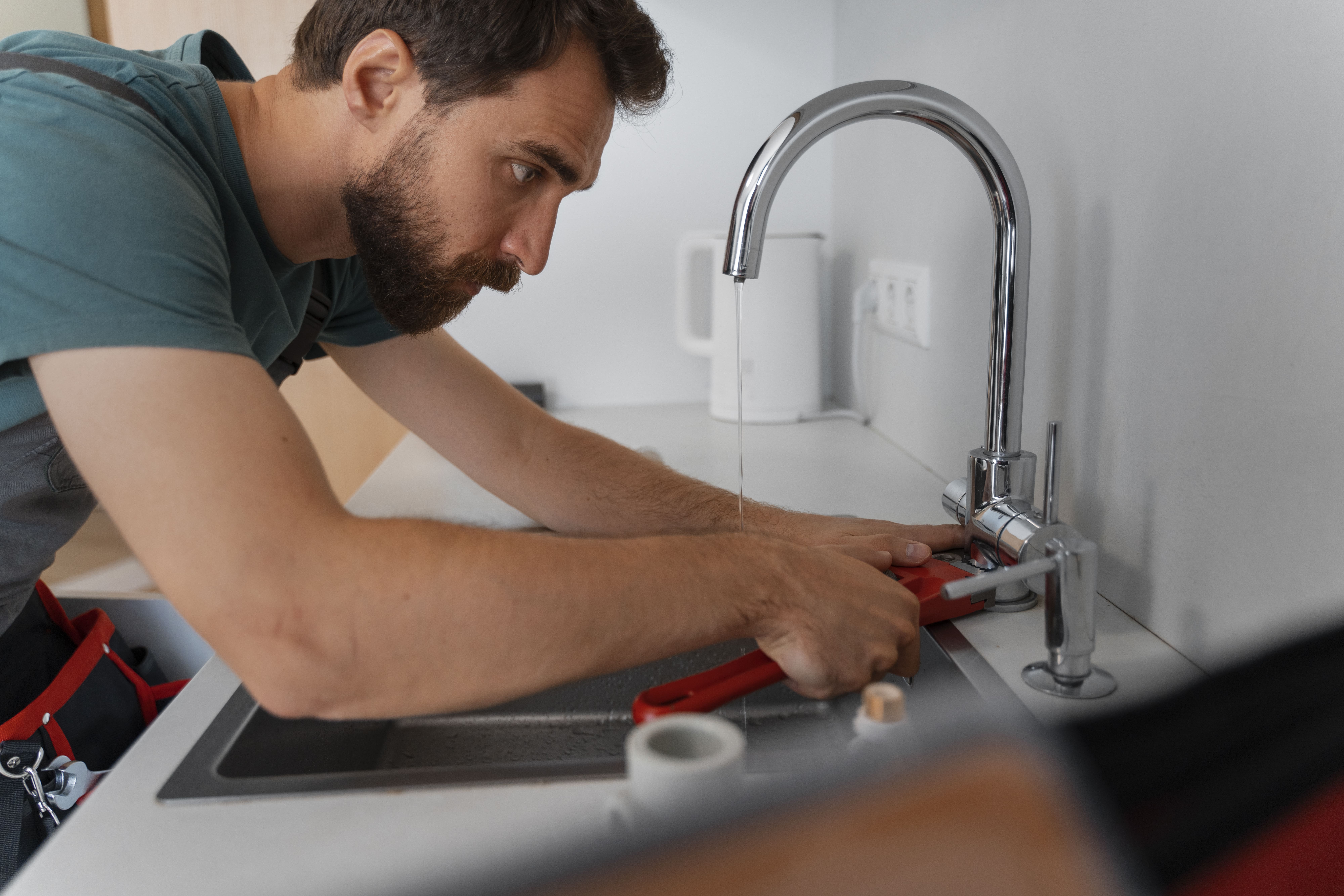 Bearded man using a red pipe wrench to fix a kitchen sink faucet with water running.