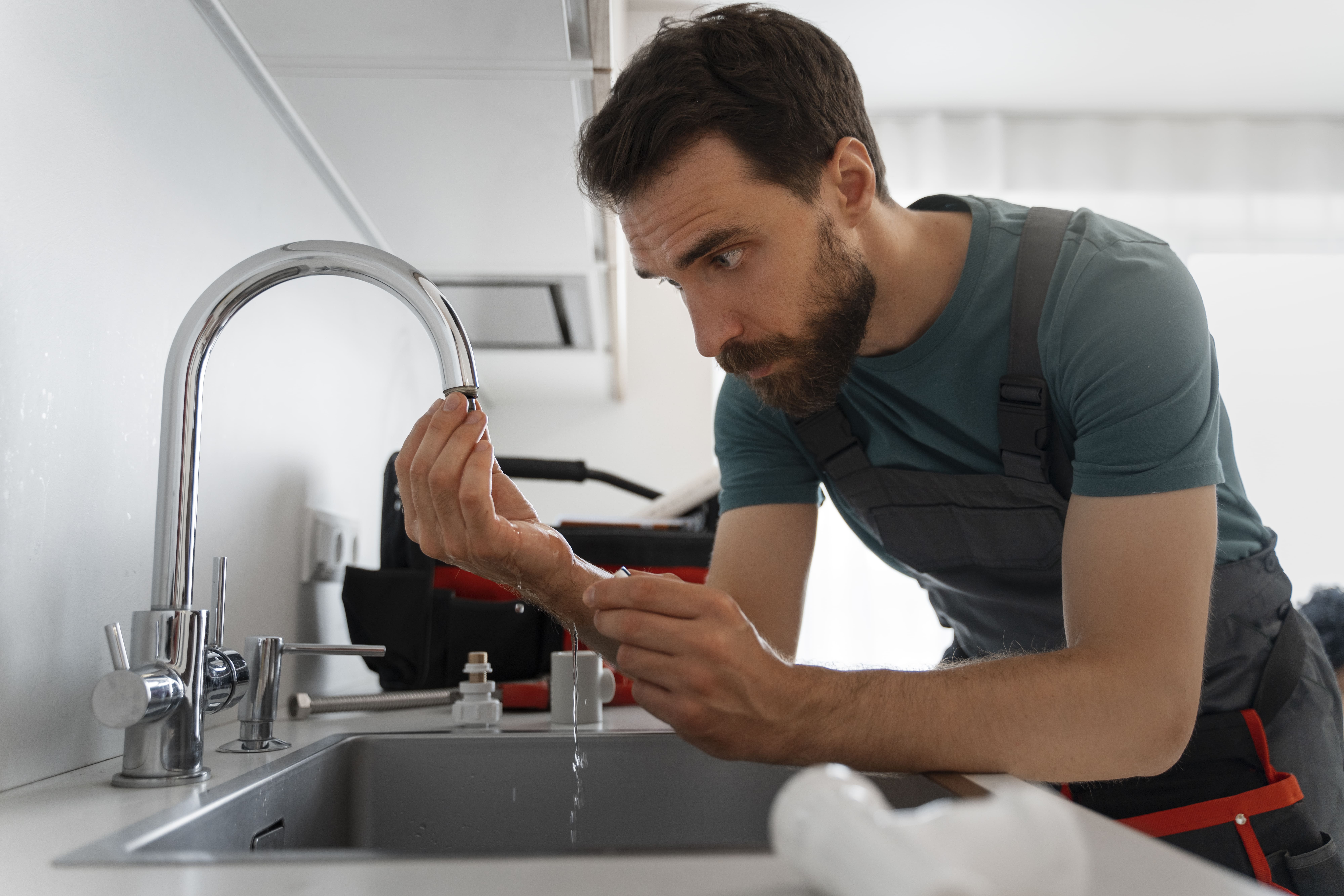 Man with a beard wearing work overalls inspecting water flow from a kitchen sink faucet.