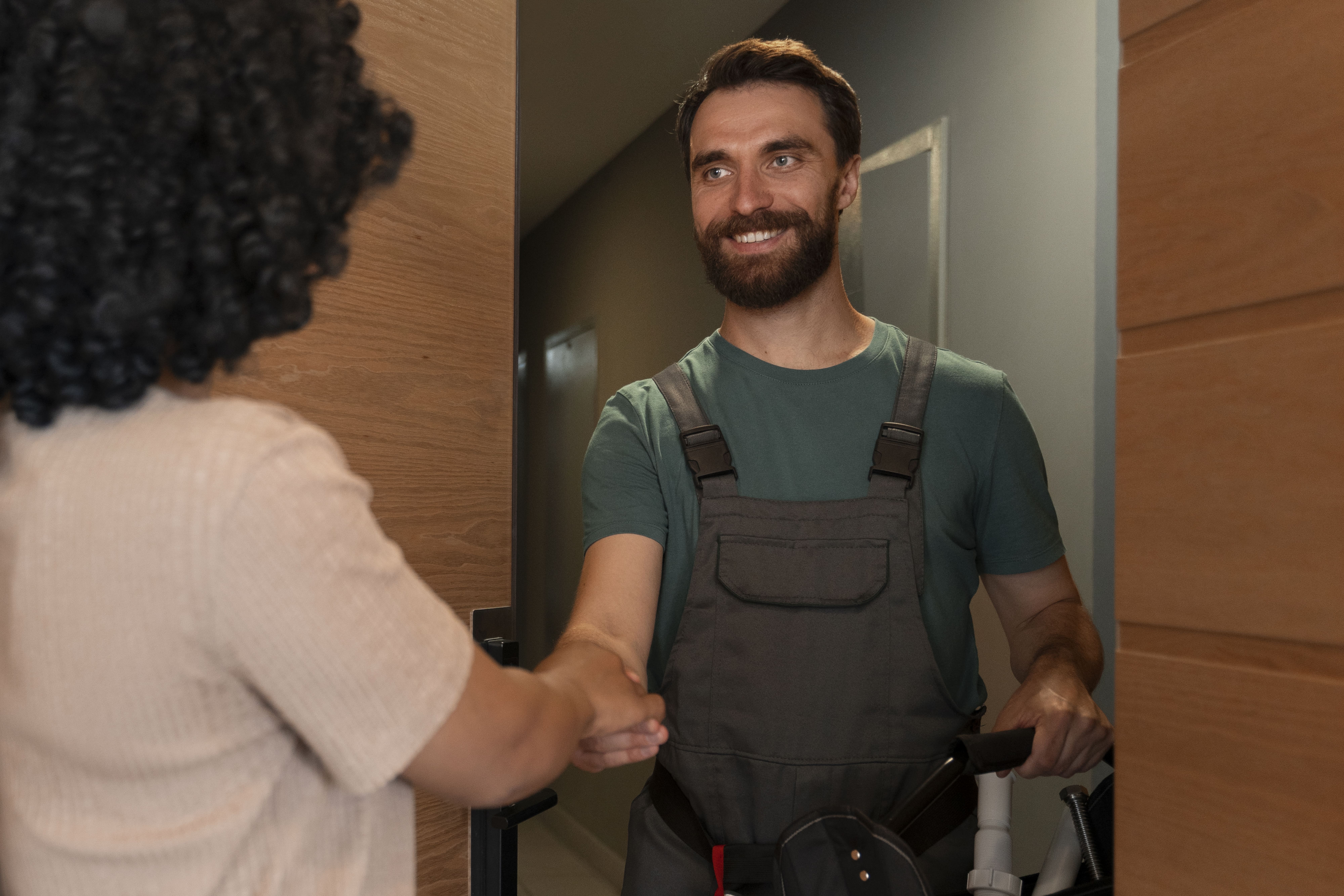 Smiling plumber in green shirt and overalls shaking hands with a woman at a door.