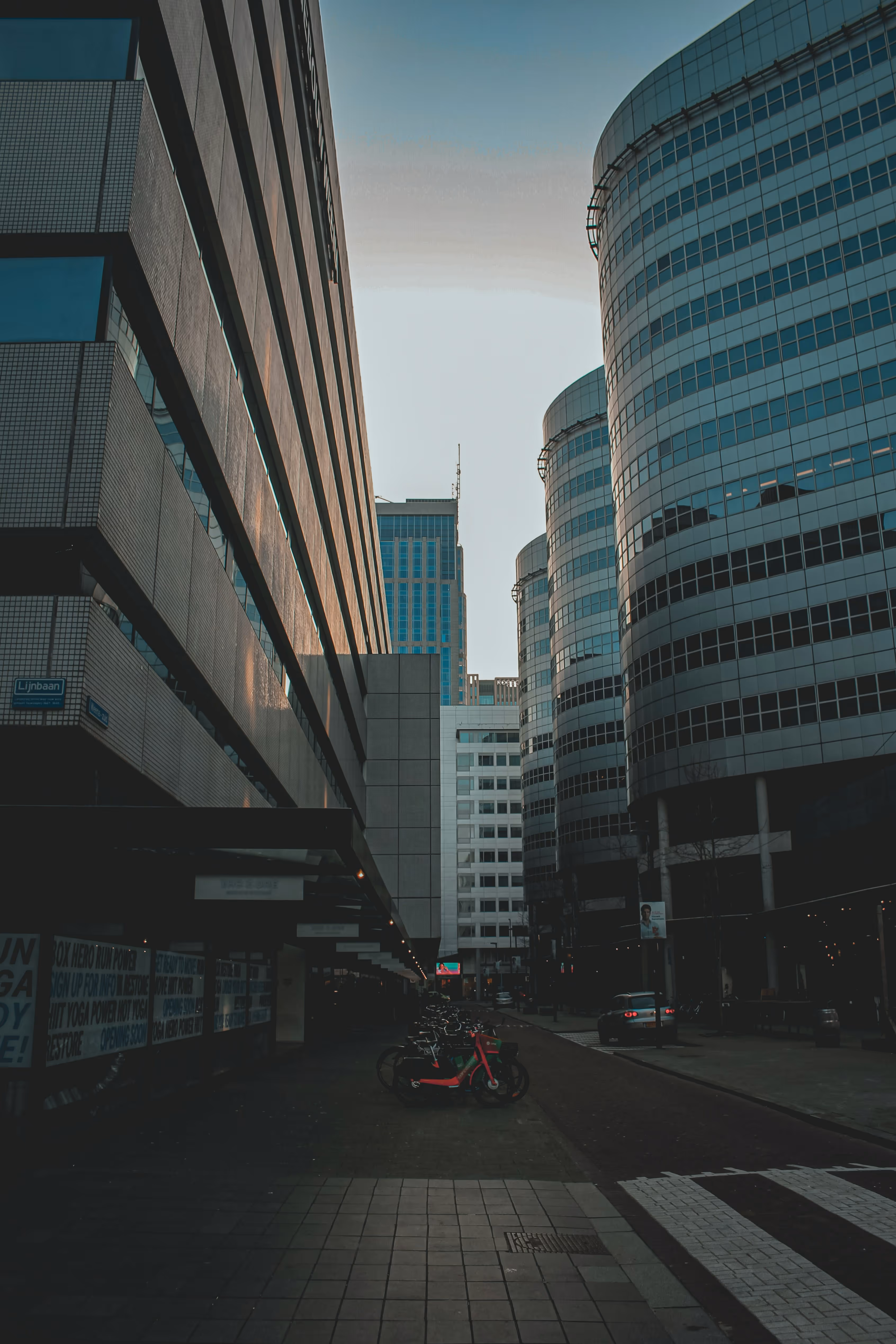 Urban street scene with modern glass and concrete buildings flanking either side and bicycles parked along the sidewalk at dusk.