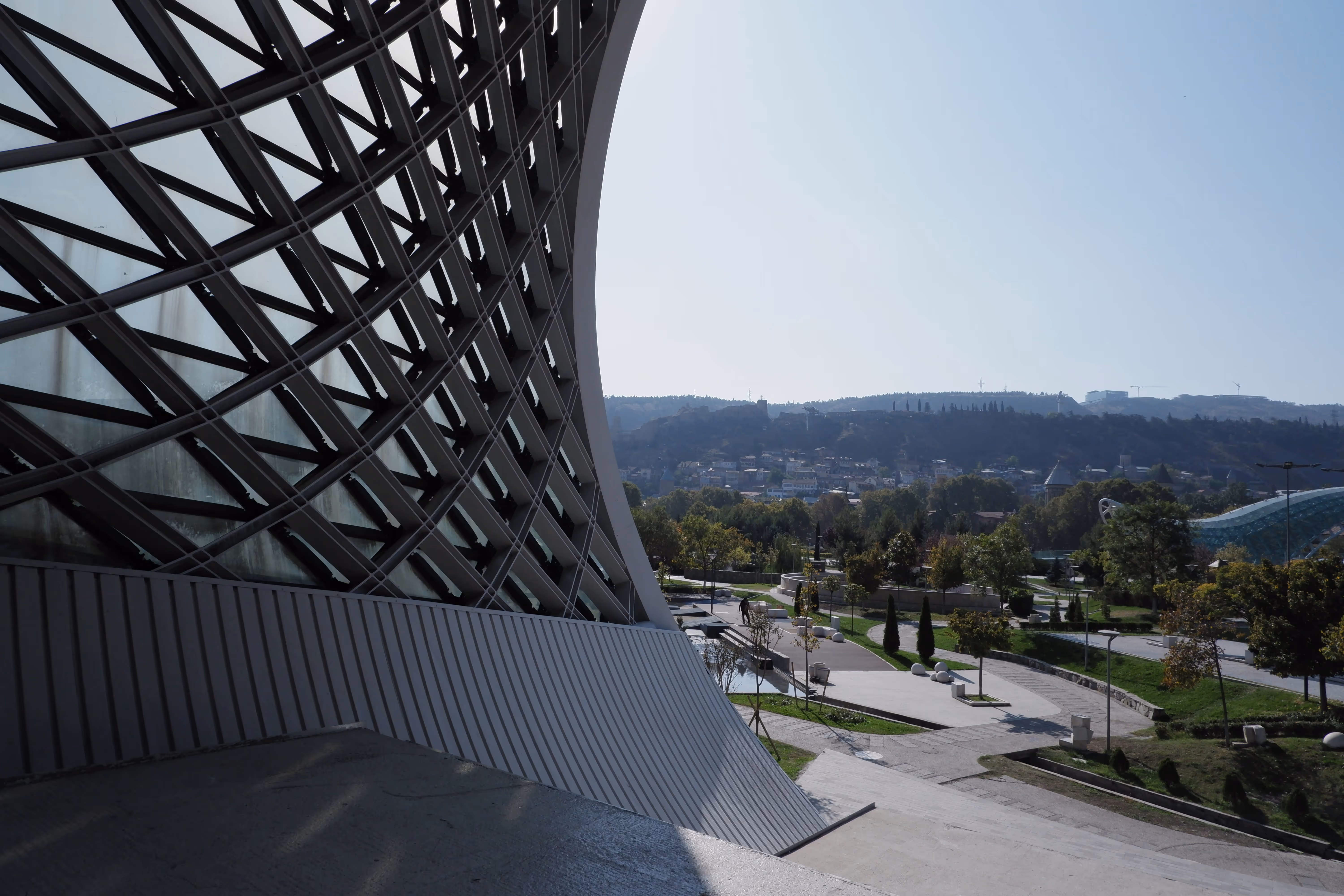 Curved modern building with large glass and steel lattice facade overlooking a park with trees, pathways, and distant hills.
