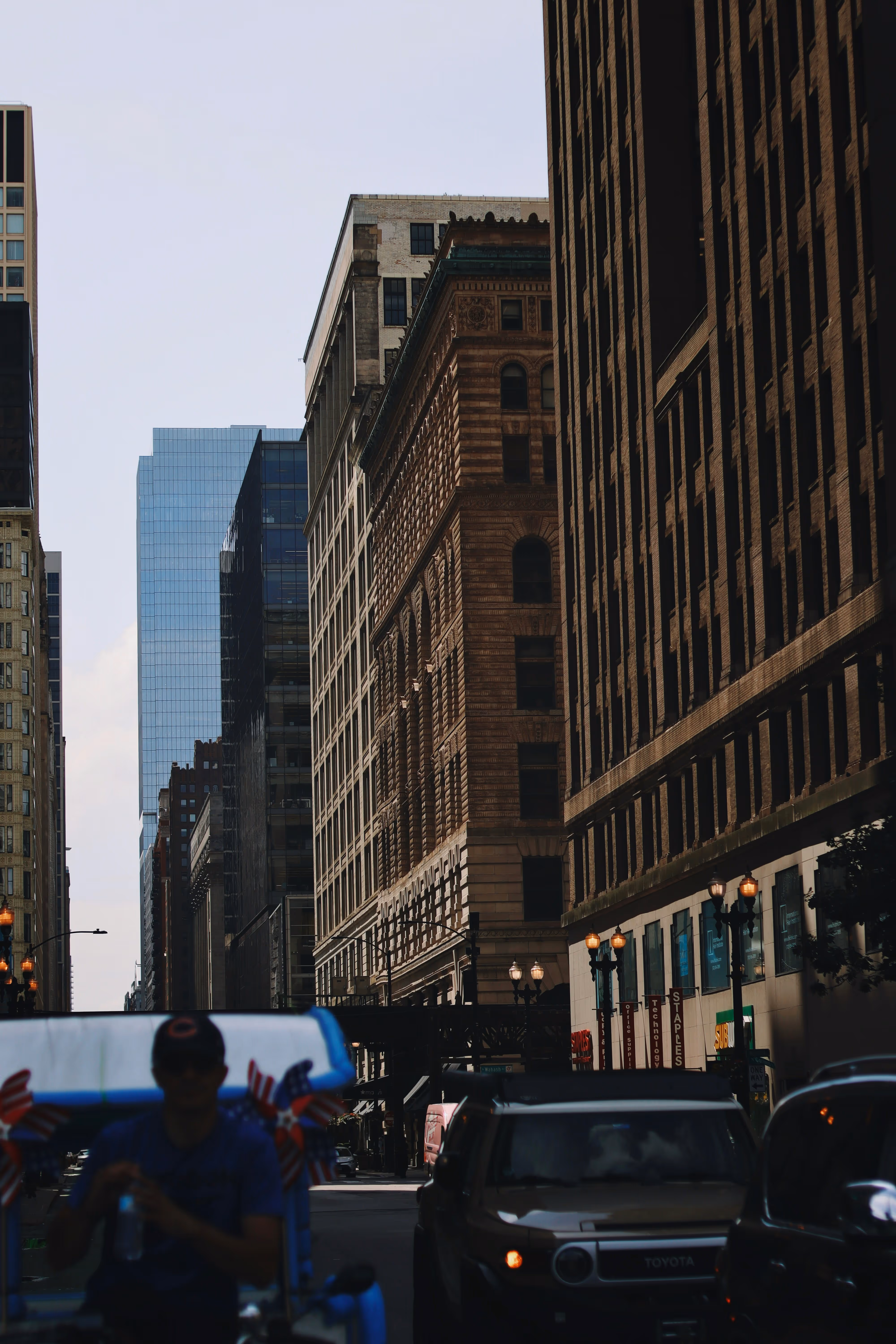 Urban street scene with tall buildings casting shadows, cars on the road, and a person riding a decorated pedicab.