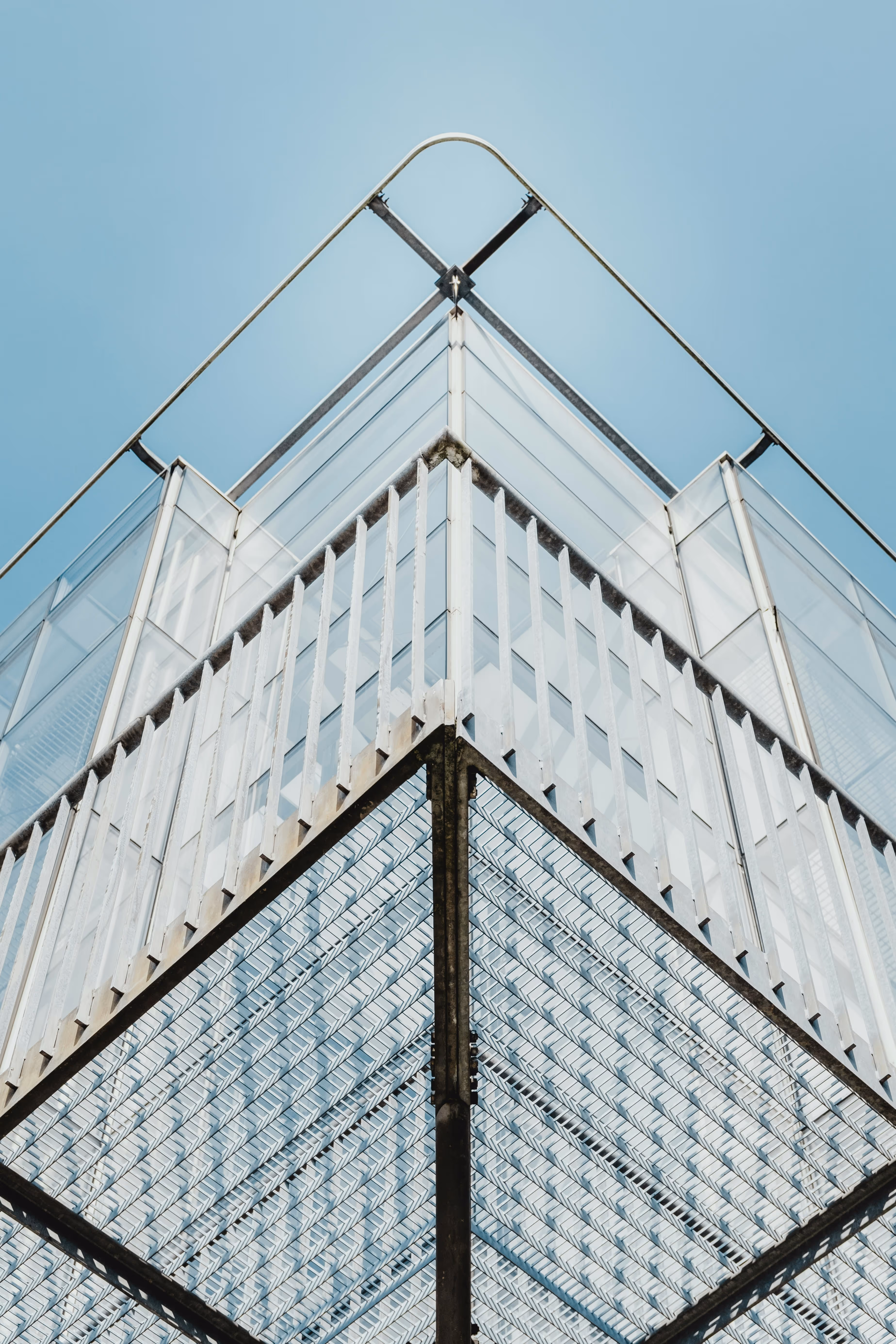 Upward view of modern building corner with glass walls and metal railing against a clear blue sky.