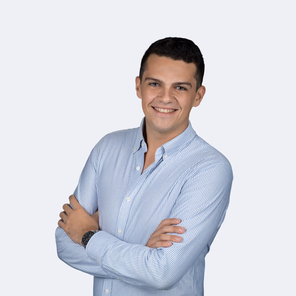 Smiling young man with short dark hair wearing a light blue striped button-up shirt with arms crossed on a light gray background.