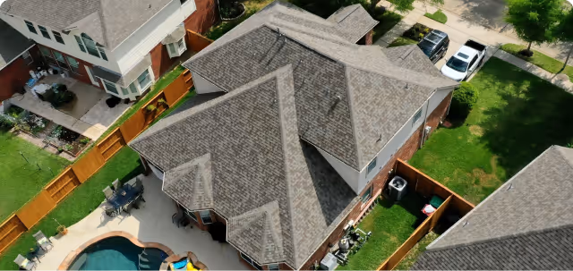 Aerial view of suburban houses with gray shingled roofs, fenced yards, and a swimming pool.