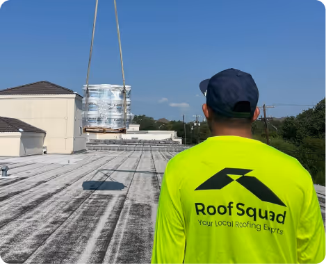 Roofer in bright yellow shirt with Roof Squad logo watches as roofing materials are lifted by crane onto a flat rooftop.