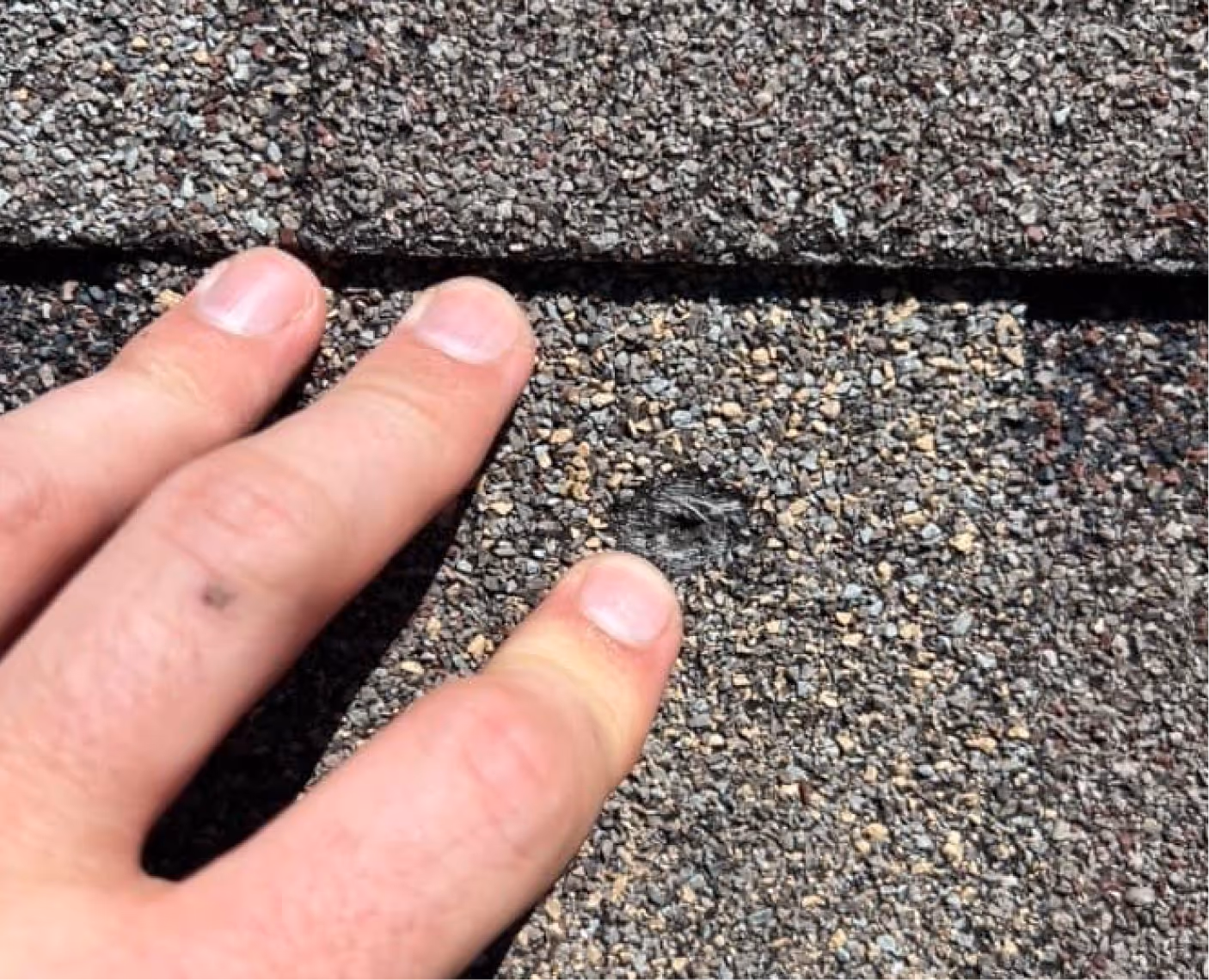 Close-up of a hand with three fingers touching a rough gray asphalt shingle roof near a visible nail hole.