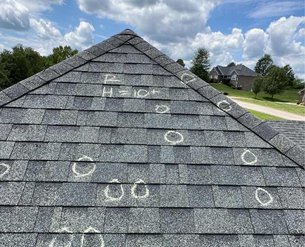 Close-up of a hand with three fingers touching a rough gray asphalt shingle roof near a visible nail hole.