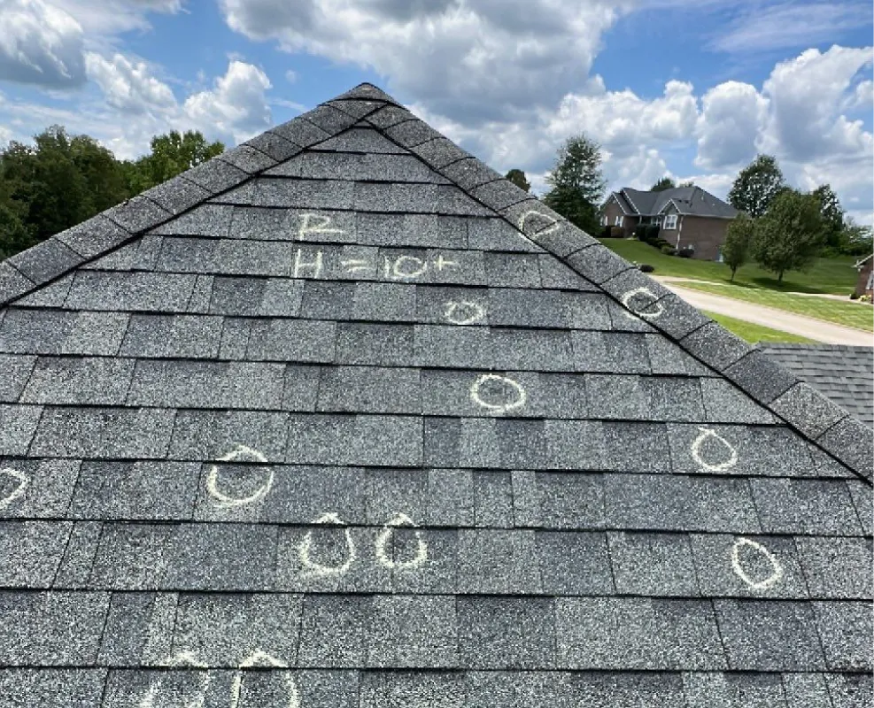 Close-up of a hand with three fingers touching a rough gray asphalt shingle roof near a visible nail hole.