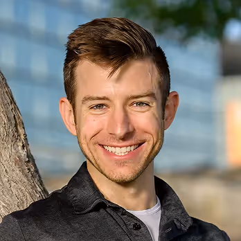 Young man with short brown hair smiling outdoors near a tree.
