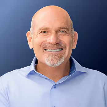Smiling bald man with light facial hair wearing a light blue collared shirt against a dark blue background.