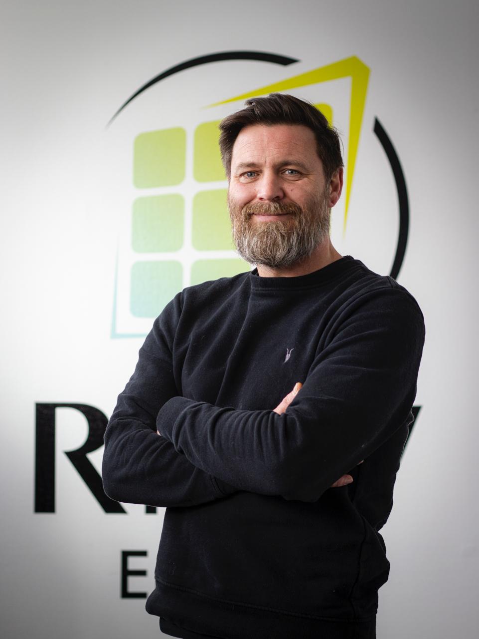 Mann mit dunklem Pullover und Bart lächelt mit verschränkten Armen vor einer Wand mit einem Logo.