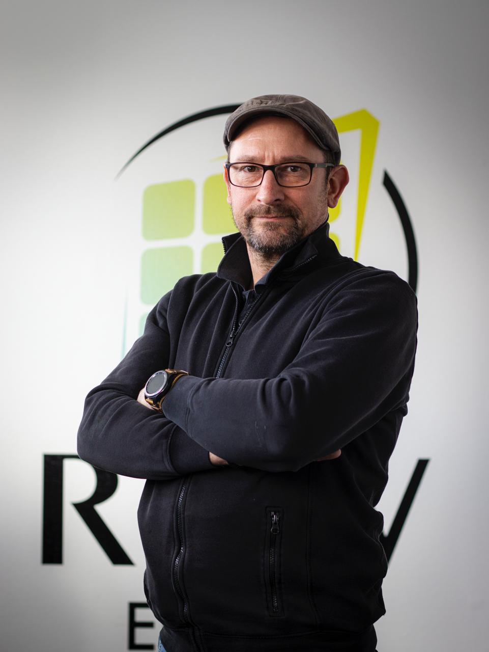 Mann mit Brille und grauer Kappe, der verschränkte Arme trägt, vor einer weißen Wand mit einem Logo.