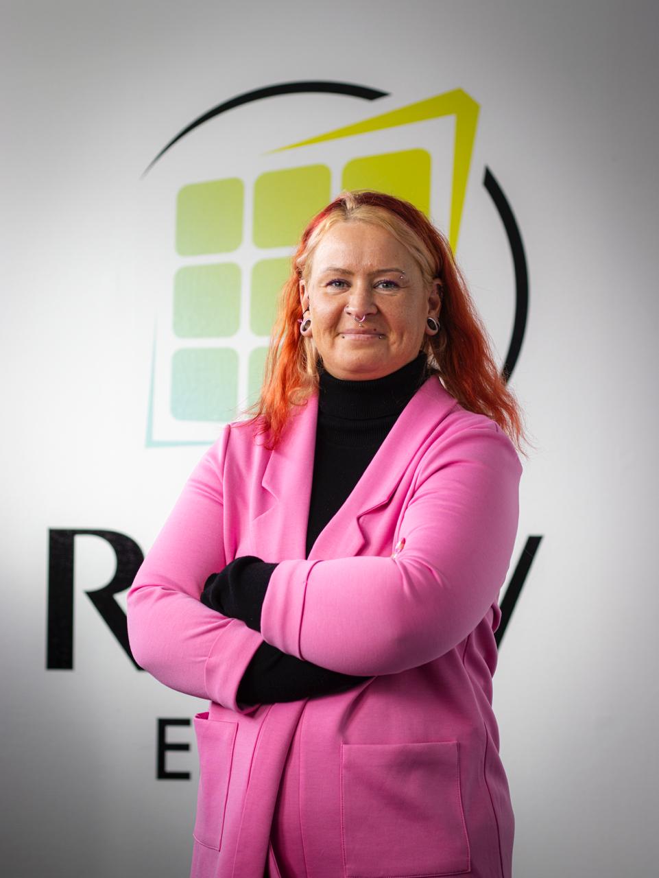 Frau mit roten Haaren, die ein pinkes Sakko und einen schwarzen Rollkragenpullover trägt, steht mit verschränkten Armen vor einer Logo-Wand.