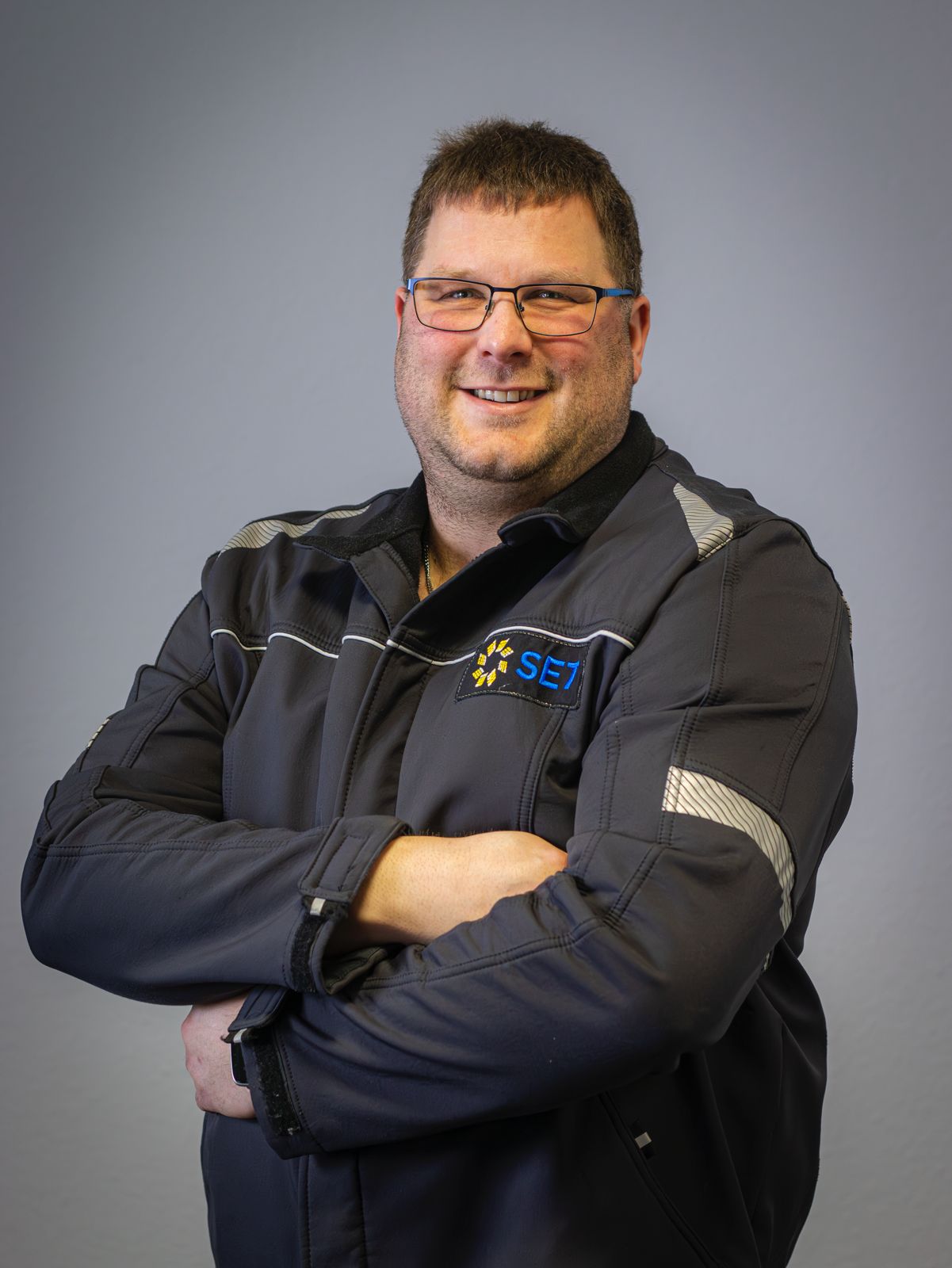 Lächelnder Mann mit Brille und dunkler Arbeitsjacke mit SET-Logo vor grauem Hintergrund.