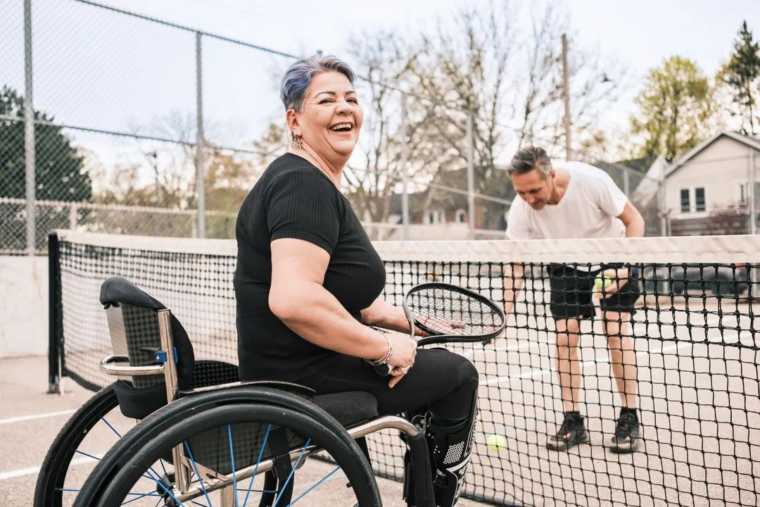 A person laughing in their wheel chair playing tennis against an abled person on a tennis court.