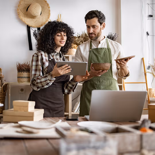 Two artisan shopowners wearing aprons reviewing a tablet and holding wooden items in a rustic workspace.