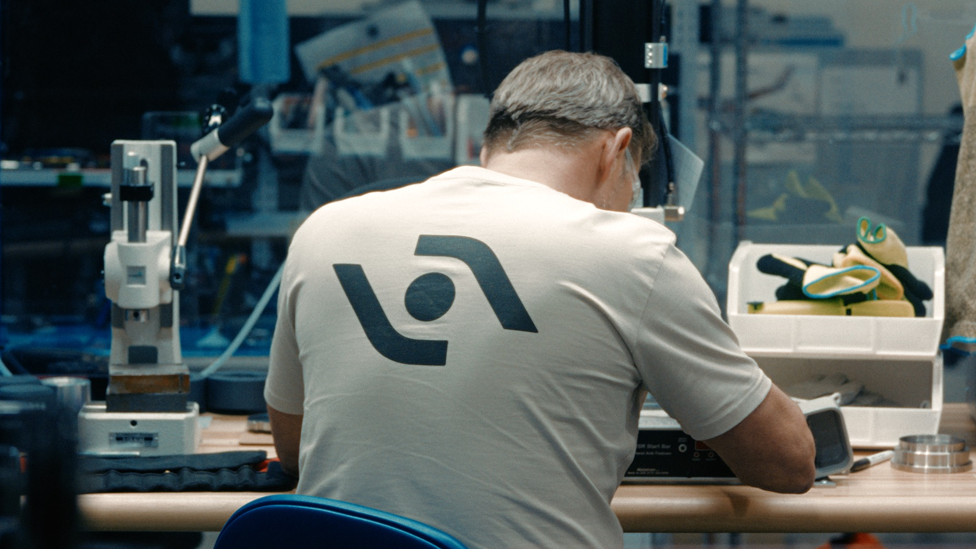 A man in a beige shirt with a black Agility logo on it sits at a work table while wearing safety goggles.