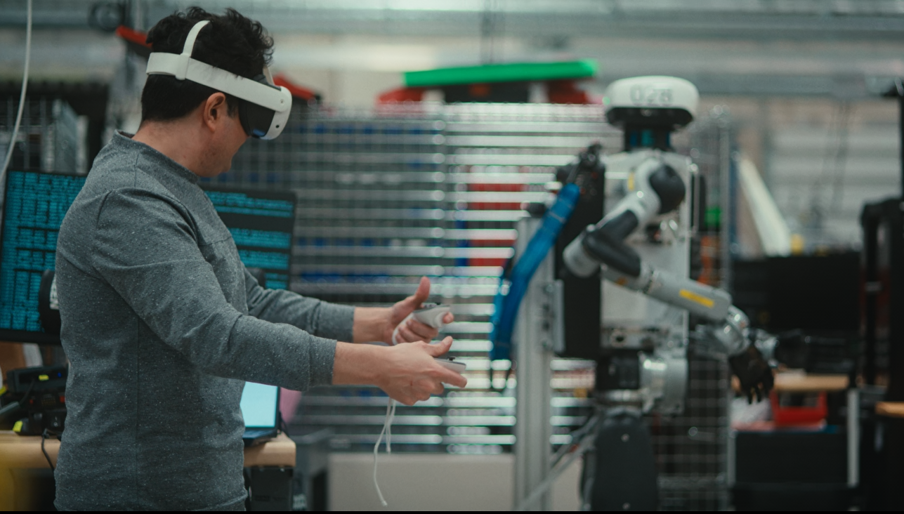 A man wearing a VR headset and holding handhold controls, with a humanoid robot in the background.
