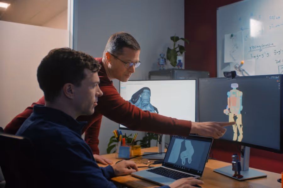 Two men working at a desk with multiple screens displaying 3D models of robotic parts.