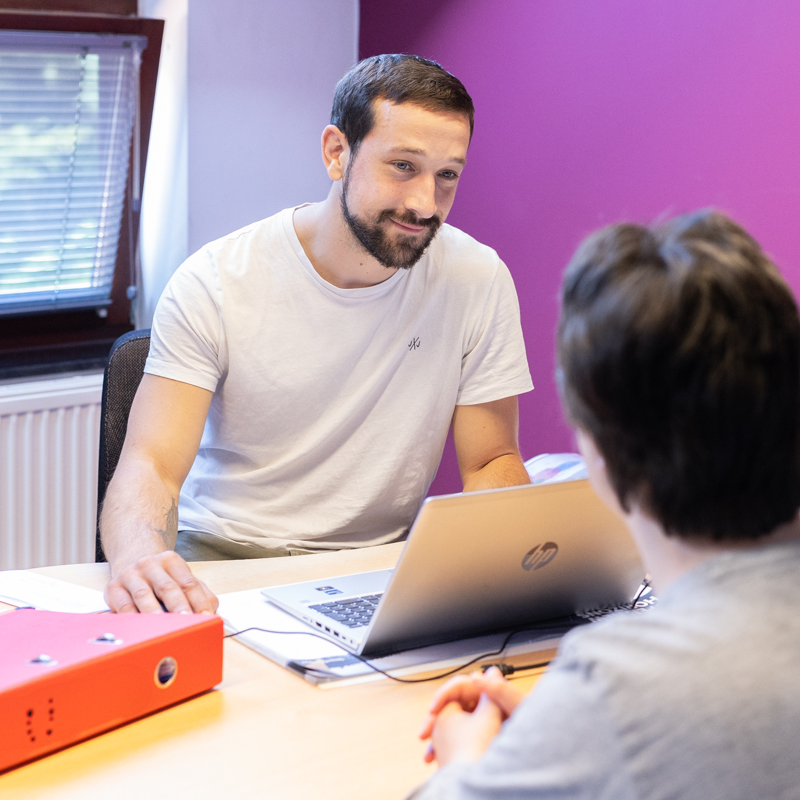 Un homme assis à un bureau en train de discuter avec une personne dont on ne voit que le dos. L'homme porte un t-shirt beige clair et semble être dans un environnement professionnel ou administratif avec des documents sur le bureau. La scène représente probablement une consultation, un entretien ou une réunion de travail.