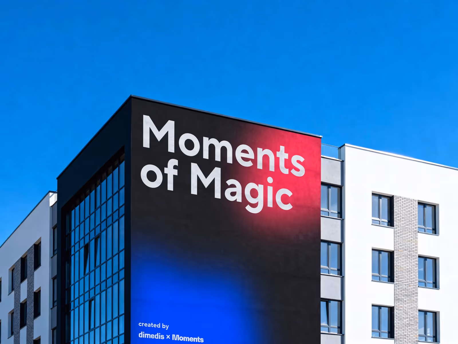 Gebäudefassade mit großem Schriftzug 'Moments of Magic' auf schwarzem Hintergrund mit roten und blauen Farbverläufen, bei klarem blauem Himmel.