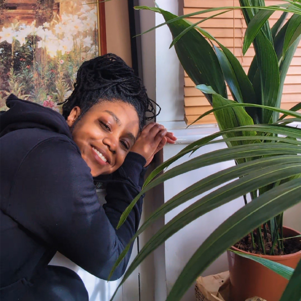 Kei is leaning on the windowsill smiling at the camera with Kentia palm leaves and an aspidistra plant next to her.