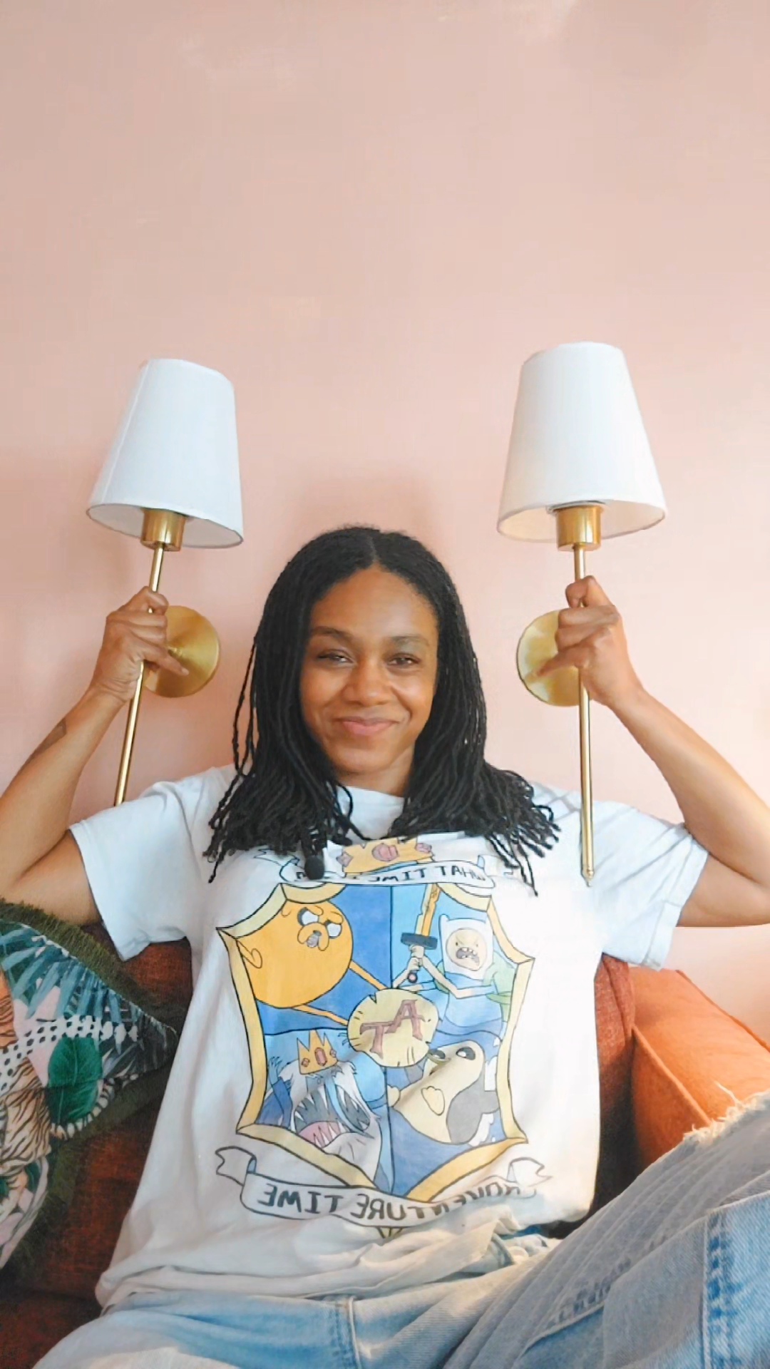 Kei is smiling while holding up two wall lampshades. She is wearing an adventure time t-shirt with stonewash jeans while sat on a red sofa.