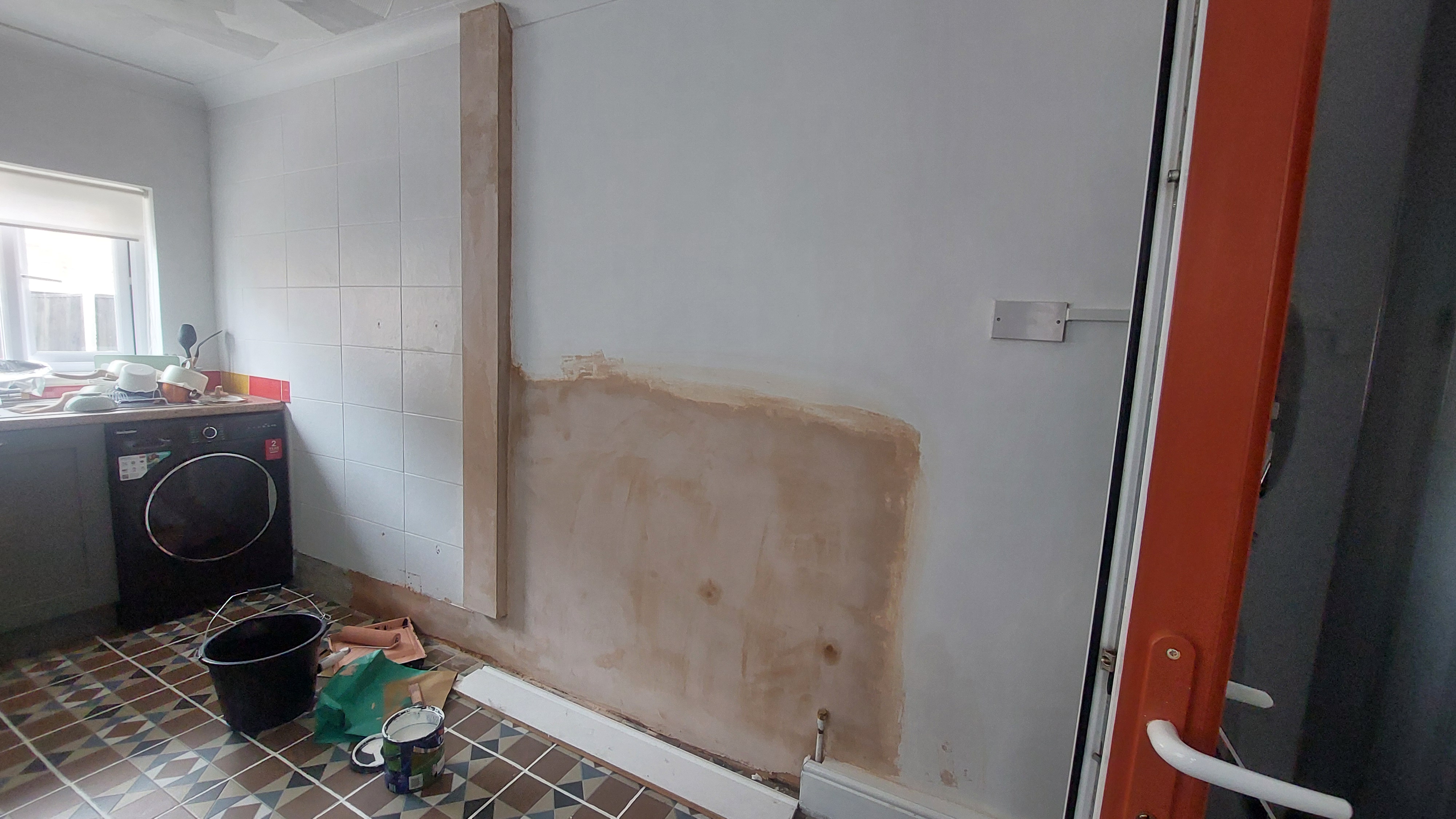 Freshly plastered kitchen wall in progress showing damp patches, patterned floor tiles, and painting supplies.