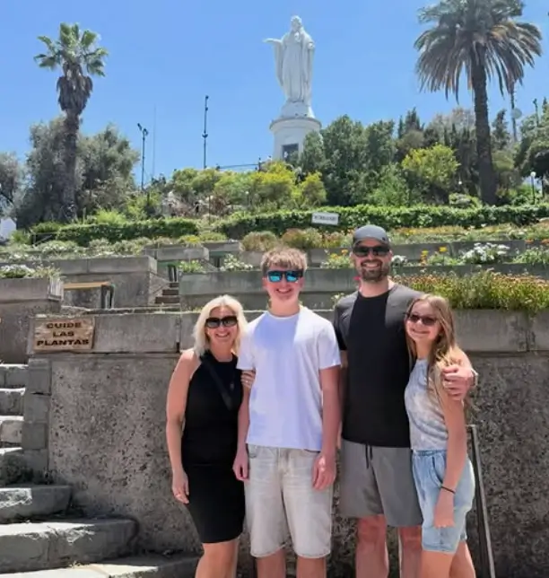 Family of four posing in front of a large statue of Jesus on a hill with palm trees and greenery on a sunny day.