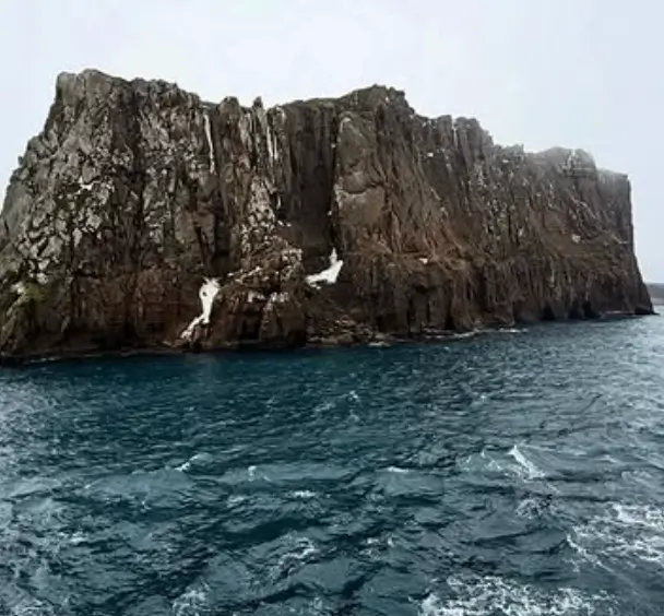 Large jagged rocky cliff rising from choppy dark blue ocean waters under a pale sky.