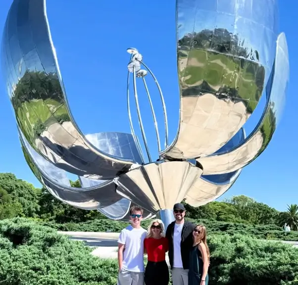 Four people standing in front of a large, shiny metallic flower sculpture outdoors on a sunny day.