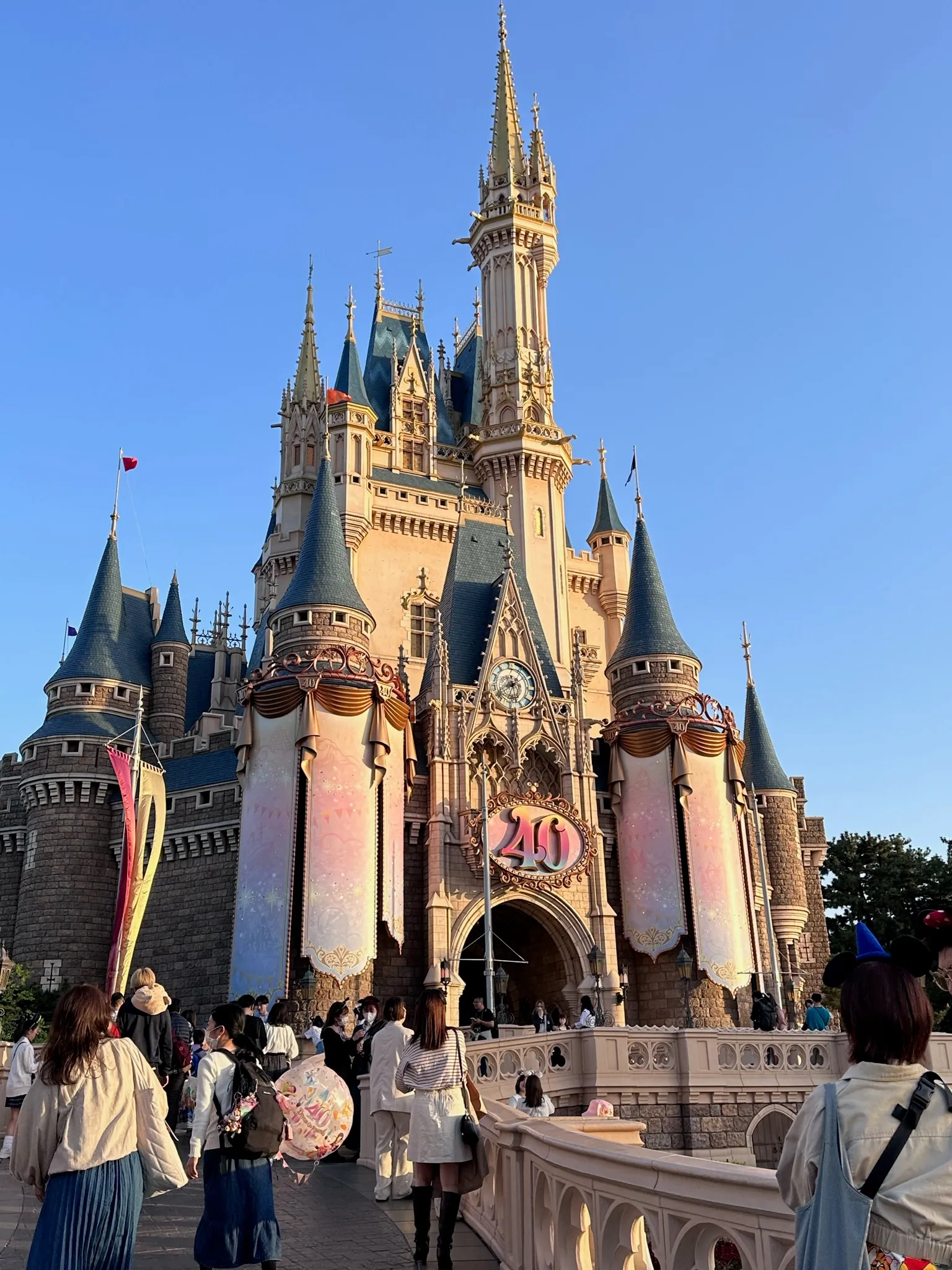 Disney castle decorated with banners displaying the number 40, with visitors walking nearby under a clear blue sky.