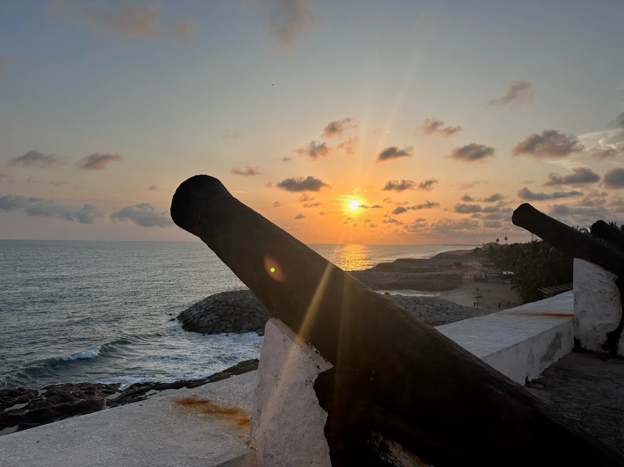 Historic cannons on a coastal fort wall overlooking the ocean at sunset with scattered clouds.