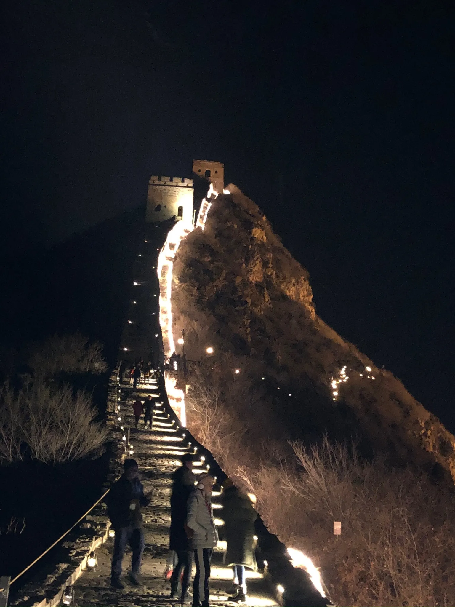 People walking on the illuminated Great Wall of China at night with watchtowers visible in the background.